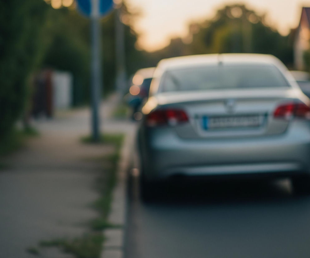 Na pierwszym planie, centralnie, widoczny jest srebrny samochód osobowy, widziany od tyłu. Pojazd stoi na poboczu drogi, z częścią karoserii wystającą na szary asfalt ulicy. Po lewej stronie samochodu rozciąga się chodnik z wyraźnymi zielonymi pasami trawy, a dalej znajduje się wysoki żywopłot oraz słup ze znakiem drogowym o okrągłym, niebieskim kształcie. Tło jest rozmyte, z widocznymi, zamazanymi kształtami innych pojazdów i zabudowań oraz ciepłym, pomarańczowo-żółtym światłem zachodzącego słońca.