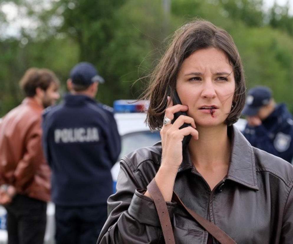 Amanda Mincewicz jako Agnes w serialu „M jak miłość”, dzwoniąca z rozciętą wargą po napaści, z Pawłem Izdebskim jako Martinem w kajdankach i policjantem na tle radiowozu. Więcej o losach bohaterów na Super Seriale.