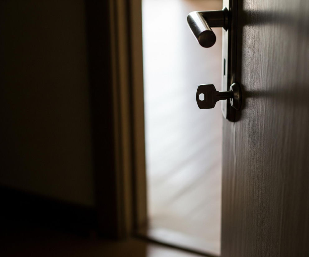 Półotwarte drewniane drzwi z metalową klamką i kluczem w zamku zajmują prawą część kadru. Klucz jest wyraźnie widoczny, wystając poziomo z zamka, z charakterystycznym otworem w swojej główce. Drzwi są ciemnobrązowe, a światło pada na ich powierzchnię, tworząc delikatne cienie i podkreślając teksturę drewna. Pomiędzy drzwiami a ciemną przestrzenią po lewej stronie widać jasny, rozmazany obszar, sugerujący światło wpadające z sąsiedniego pomieszczenia, odbijające się od podłogi. Lewa strona obrazu pozostaje w głębokim cieniu, tworząc kontrast z oświetlonym przejściem.