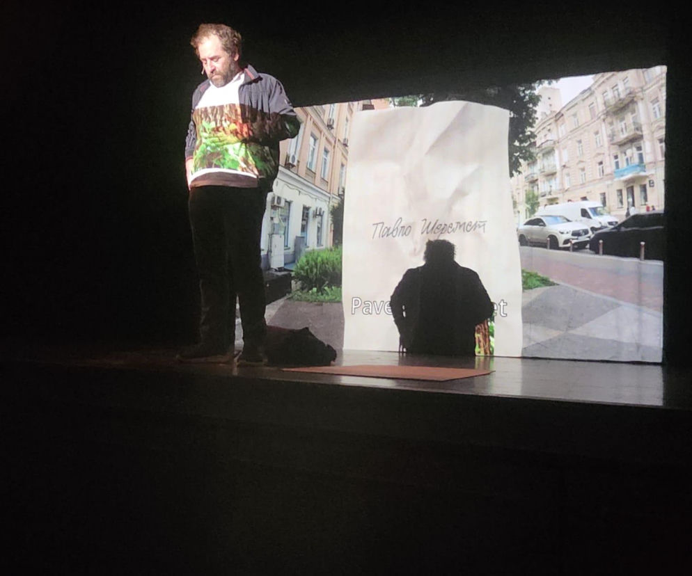 Aktor Piotr Armianowski na scenie podczas spektaklu If I Had a Gun, I'd Take Them All Down w Bohemian National Hall, z projekcją ulic Kijowa w tle, poruszającego temat wojny w Ukrainie. Więcej o przedstawieniu przeczytasz na naszym portalu.