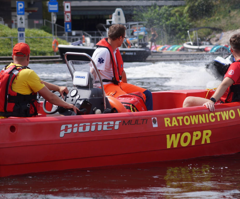 Czerwona łódź ratownictwa wodnego WOPR z trzema ratownikami w kamizelkach ratunkowych na pokładzie, płynąca po wodzie, za nimi spieniona woda i inne łodzie. Zdjęcie ilustruje akcję ratunkową i apel o ostrożność nad wodą, o czym przeczytasz na naszym portalu.