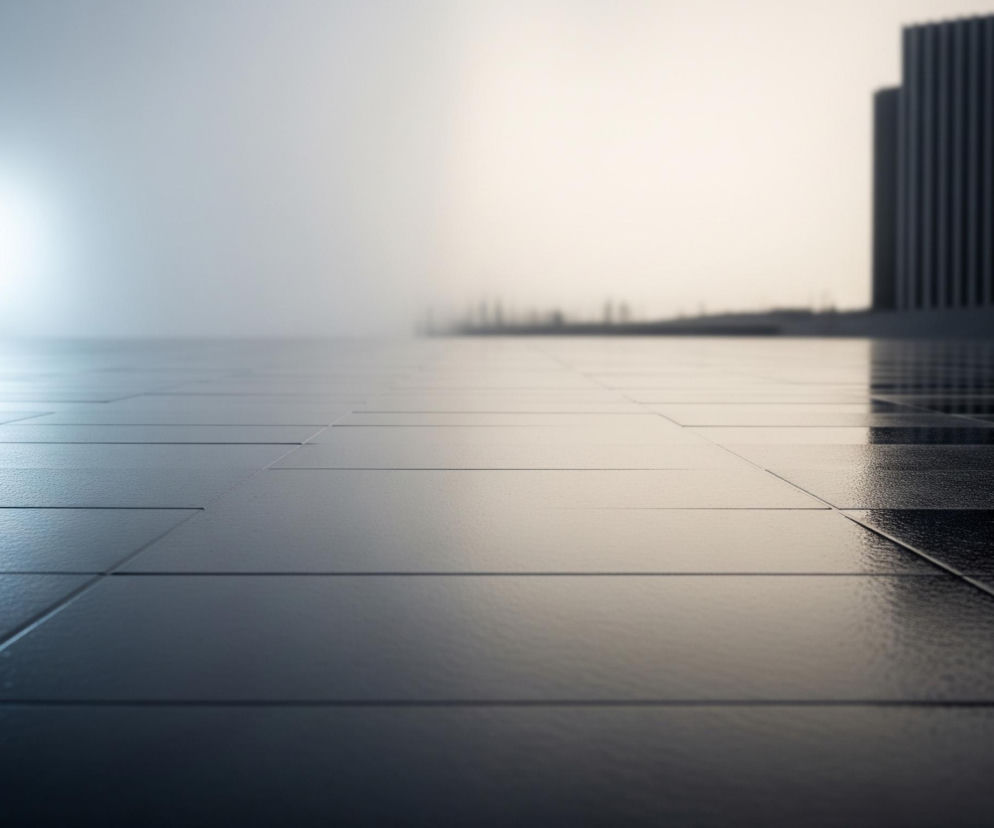 Płaskie, lśniące, ciemne płytki pokrywają cały pierwszy plan i rozciągają się w głąb obrazu. Po lewej stronie dominuje jasne, białe światło, tworzące odblaski na powierzchni, podczas gdy prawa strona jest cieplejsza, o beżowo-żółtawym odcieniu. Na horyzoncie widoczne są niewyraźne, ciemne sylwetki budynków, w tym jedna wyraźnie większa, o pionowych liniach, przypominająca wieżowiec. Całość sprawia wrażenie minimalistycznego, miejskiego pejzażu, w którym dominują gładkie tekstury i gradienty świetlne.