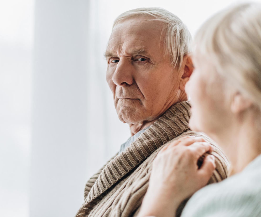 Starszy mężczyzna z zatroskaną miną patrzy prosto w obiektyw, gdy młodsza kobieta kładzie mu dłoń na ramieniu, symbolizując wsparcie w chorobie. Więcej o anemii i demencji, przeczytasz na portalu Poradnik Zdrowie.