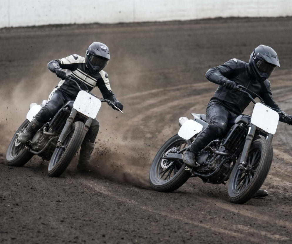 Dwóch żużlowców w pełnych kombinezonach i kaskach ściga się na motocyklach na torze, wzbijając tumany kurzu. Emocjonująca scena wyścigu ligowego, o której więcej przeczytasz na naszym portalu.