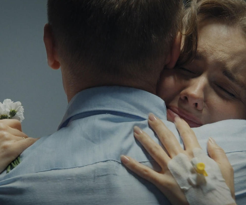Uścisk Franki (Dominika Kachlik) i Pawła (Rafał Mroczek) w szpitalu, Franka płacze, a na jej ręce widoczny jest wenflon, co potęguje dramatyzm sceny. W ręce trzyma białe kwiaty, symbol nadziei lub pożegnania. Więcej o ich losach na Super Seriale.