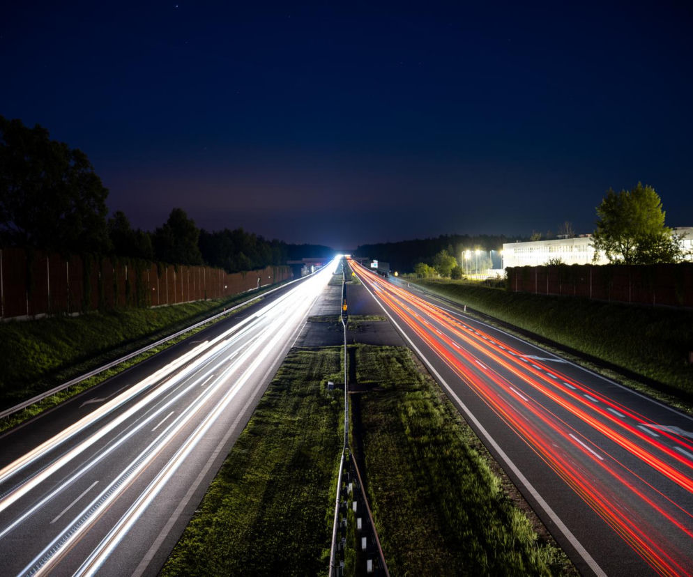 Nocny widok na oświetloną autostradę A4 Katowice-Kraków z smugami świateł pojazdów, symbolizujący ruch i zmiany stawek opłat, o których można przeczytać na Super Biznes. Po obu stronach widoczne ekrany akustyczne i zieleń.