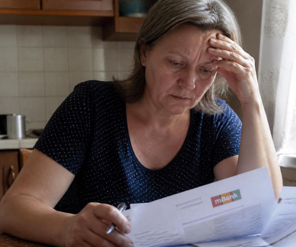 Zmartwiona kobieta w średnim wieku, Irena, z dłonią na czole, siedzi przy stole, analizując dokumenty finansowe z logo mBank. Zdjęcie ilustruje trudną decyzję o hipotece domu, która może zrujnować małżeństwo, o czym można przeczytać na naszym portalu.