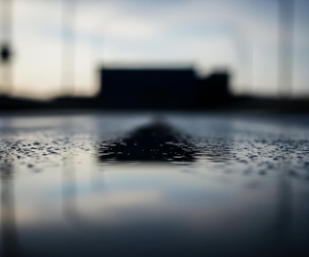 Powierzchnia pokryta licznymi drobnymi kropelkami wody znajduje się w dolnej części obrazu, rozciągając się od dołu do około jednej trzeciej wysokości kadru, gdzie staje się gładka i lustrzana. W oddali, po lewej i prawej stronie, widoczne są rozmyte, okrągłe źródła światła, odpowiednio czerwone i niebieskie, których odbicia w tych samych kolorach widać wyraźnie na mokrej powierzchni poniżej. Centralnie w tle, wyraźnie zamazana, ciemna sylwetka budynku lub innej konstrukcji unosi się na tle jaśniejszego, niewyraźnego nieba, dominując środkowy plan.