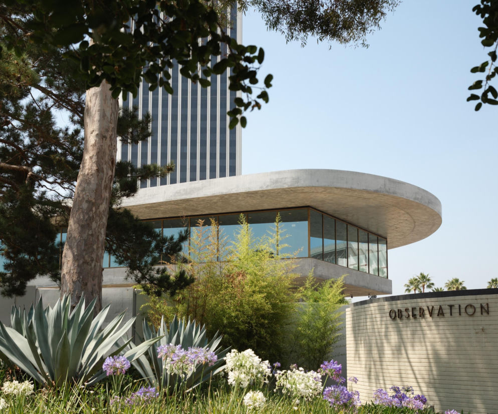 Widok na Muzeum LACMA w Los Angeles, zaprojektowane przez Petera Zumthora, z dominującą betonowo-szklaną fasadą i zaokrąglonymi kształtami, otoczone bujną, śródziemnomorską roślinnością. Więcej o architekturze tego miejsca znajdziesz na Architektura Murator Plus.