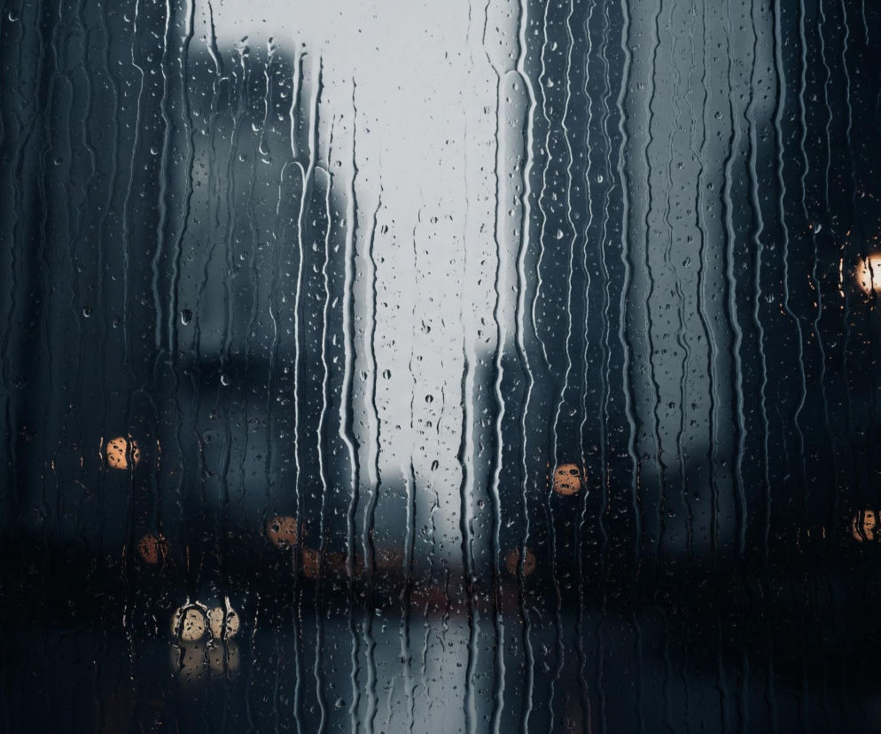 Przednia płaszczyzna obrazu to szyba pokryta licznymi kroplami deszczu, które spływają wzdłuż pionowych smug, tworząc nieregularne wzory i faliste linie. Za mokrą szybą widać zamazane kształty budynków, z których centralny jest jaśniejszy i wydaje się być odleglejszy. Na dole, w tle, widoczne są rozmyte, pomarańczowo-żółte światła miejskie, które odbijają się na wilgotnej powierzchni, tworząc efekt bokeh. Dominują ciemne odcienie szarości i błękitu, przełamane ciepłymi punktami świetlnymi.