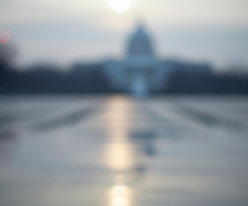 Rozmyty widok rozległego placu z dominującą w tle sylwetką budynku z kopułą. Na pierwszym planie, po lewej stronie, widoczne są rozmyte światła samochodowe w kolorach niebieskim, czerwonym i żółto-pomarańczowym. Centralna część placu odbija jasne światło, tworząc długą, świetlistą smugę prowadzącą w głąb obrazu, w kierunku budynku. Niebo jest jasne z widocznym rozbłyskiem słońca w górnej części kadru.