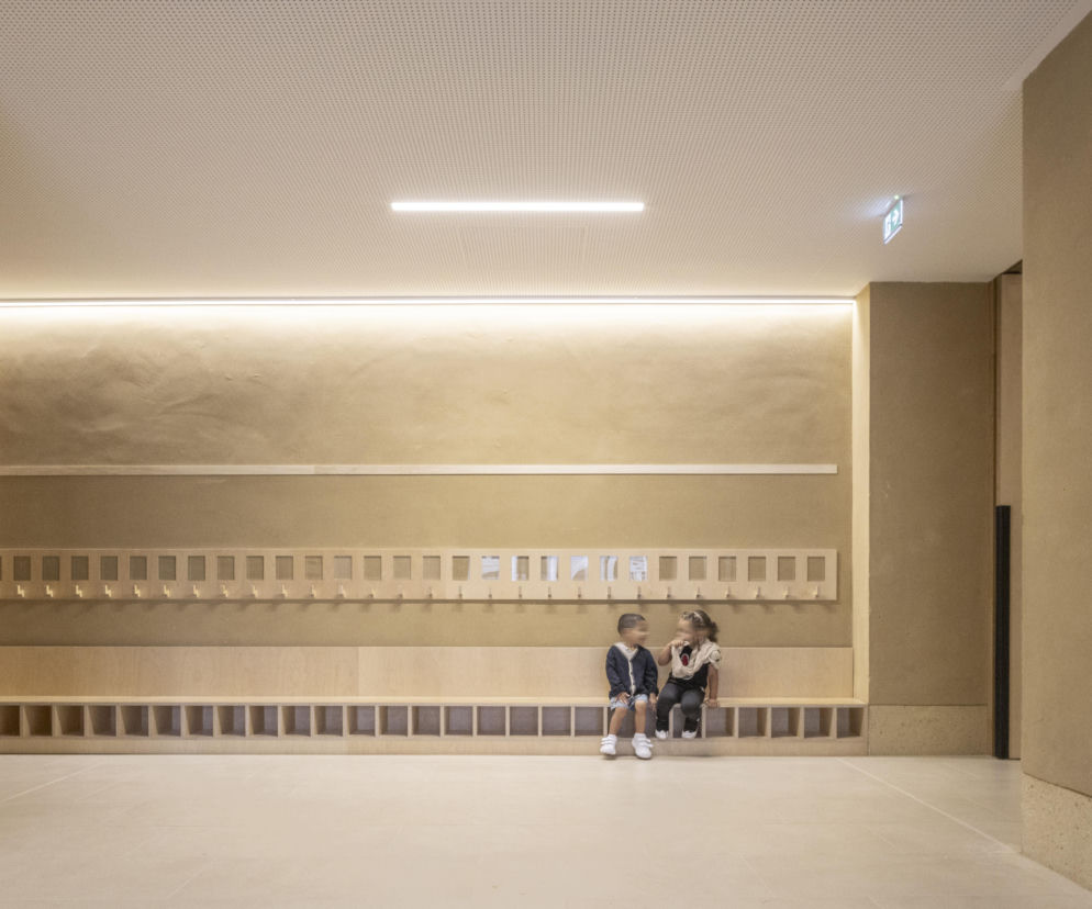 Korytarz szkoły w Châtenay-Malabry z drewnianymi panelami i beżowymi ścianami. Na ławce siedzą dwoje dzieci. Wyróżniony projekt świadczy o innowacyjnym podejściu do ekologii, o czym można przeczytać w Architektura Murator Plus.