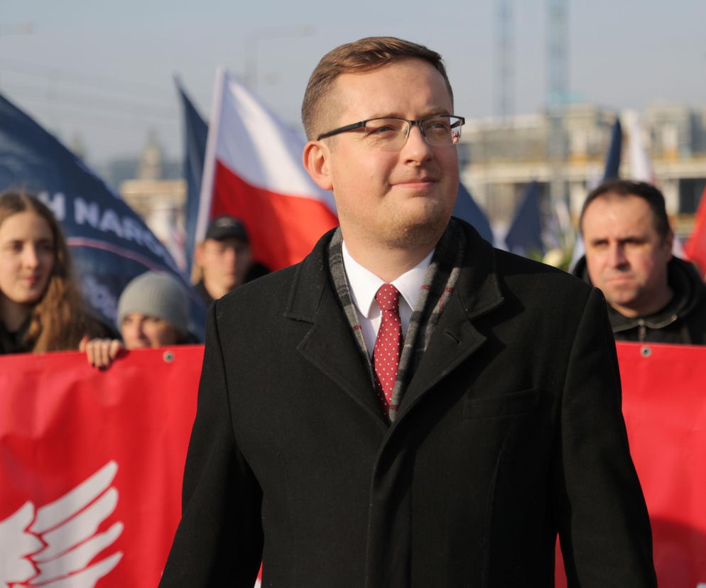 Robert Winnicki w ciemnym płaszczu na tle flag. O krytyce Trybunału Konstytucyjnego przeczytasz na SE Polityka.