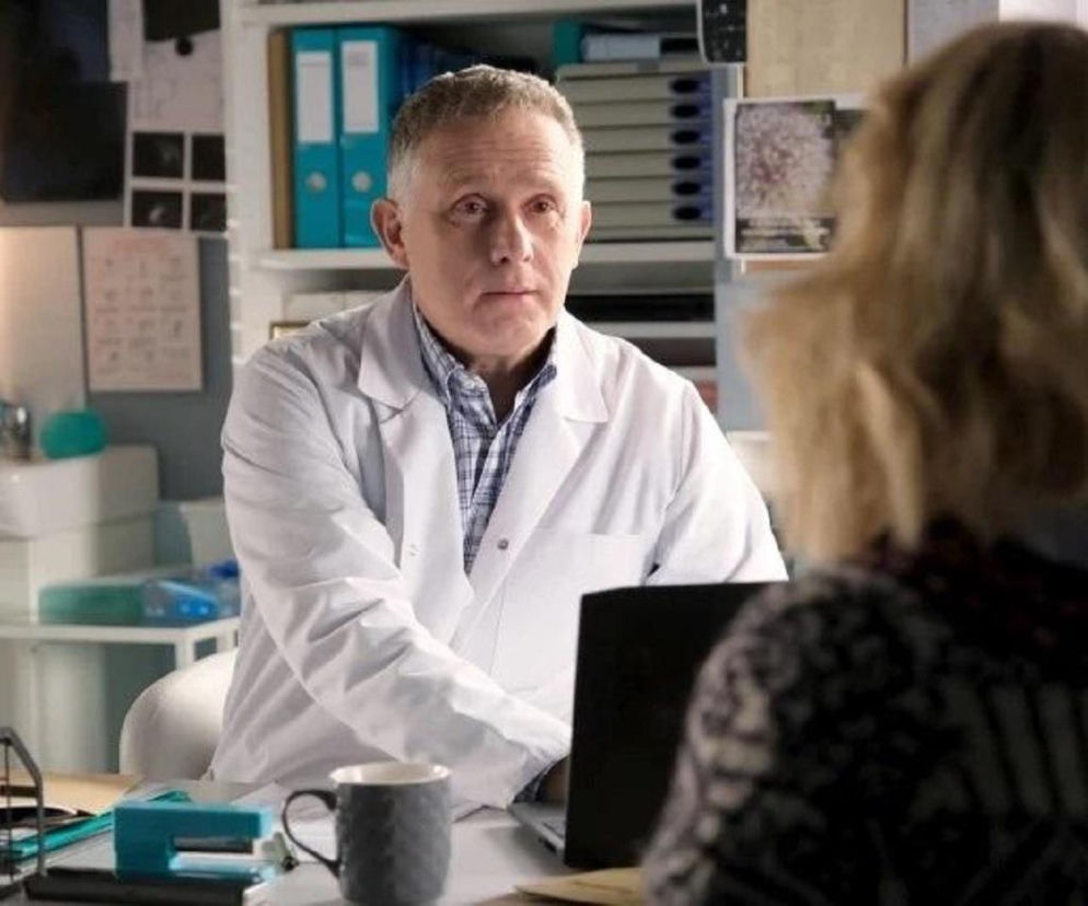 Artur Rogowski (Robert Moskwa) w białym lekarskim fartuchu, siedzący za biurkiem i rozmawiający z Judytą (Paulina Chruściel) widoczną od tyłu. Scena z serialu M jak miłość, odcinek 1931, gdzie Judyta ostrzega Artura przed zdradą Marysi. Więcej o tym na stronie Super Seriale.