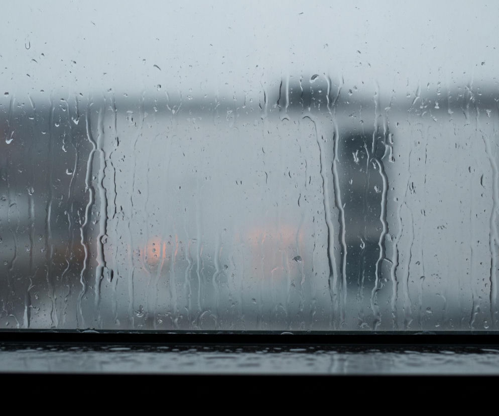 Przez szybę z licznymi kroplami deszczu spływającymi pionowo widać rozmyty, szary krajobraz miejski. W tle znajdują się niewyraźne, ciemniejsze budynki i jaśniejsze niebo. Na środku obrazu, za mokrą szybą, delikatnie prześwitują dwie pomarańczowe poświaty, przypominające światła. Dolna część kadru jest ciemna, tworząc wyraźną, poziomą linię oddzielającą.