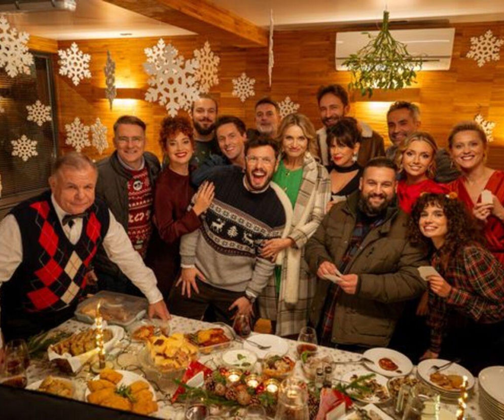 Uśmiechnięta obsada serialu Mikołaj Na sygnale 2 podczas świątecznego spotkania, ze stołem pełnym potraw i dekoracjami ze śnieżynek. Aktorzy pozują razem, tworząc radosną atmosferę, co stanowi idealne wprowadzenie do tematu świątecznych zmian w ramówce TVP2, o których możesz przeczytać na portalu Super Seriale.