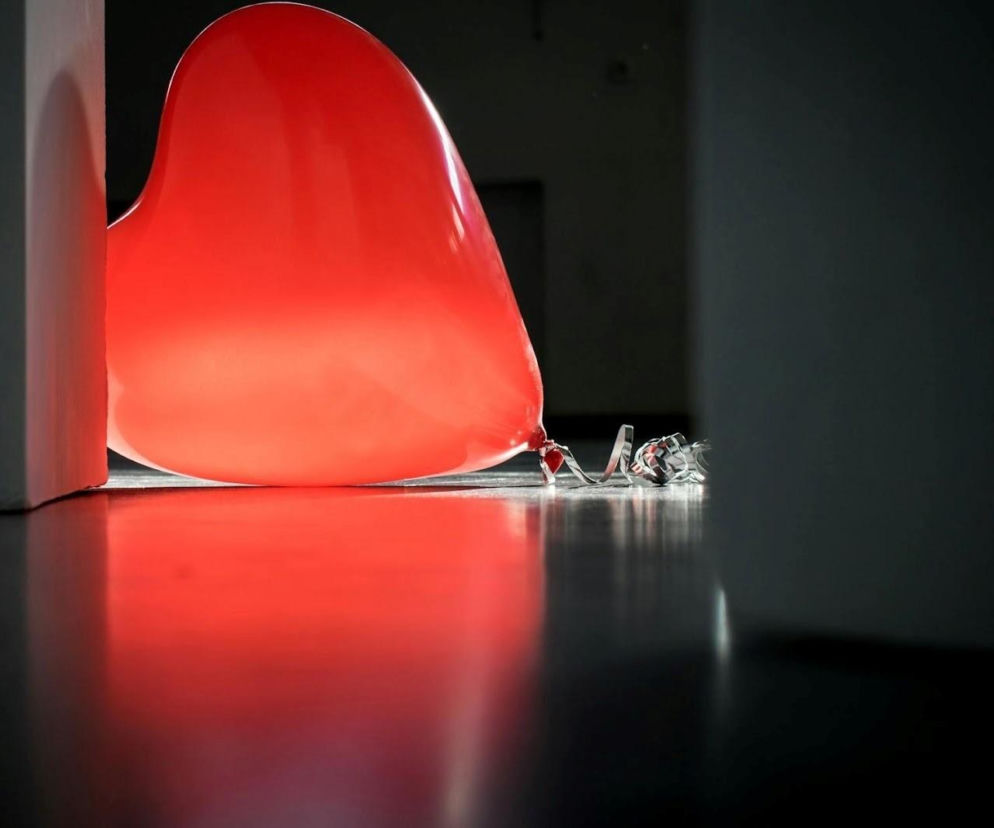 Czerwony balon w kształcie serca leżący na ciemnej, lśniącej podłodze. Obok niego, za fragmentem białej ściany, widoczne są zwinięte, srebrne serpentyny. Balon jest głównym elementem, który symbolizuje walentynki i zmysłowość, o której przeczytasz na Poradnik Zdrowie.