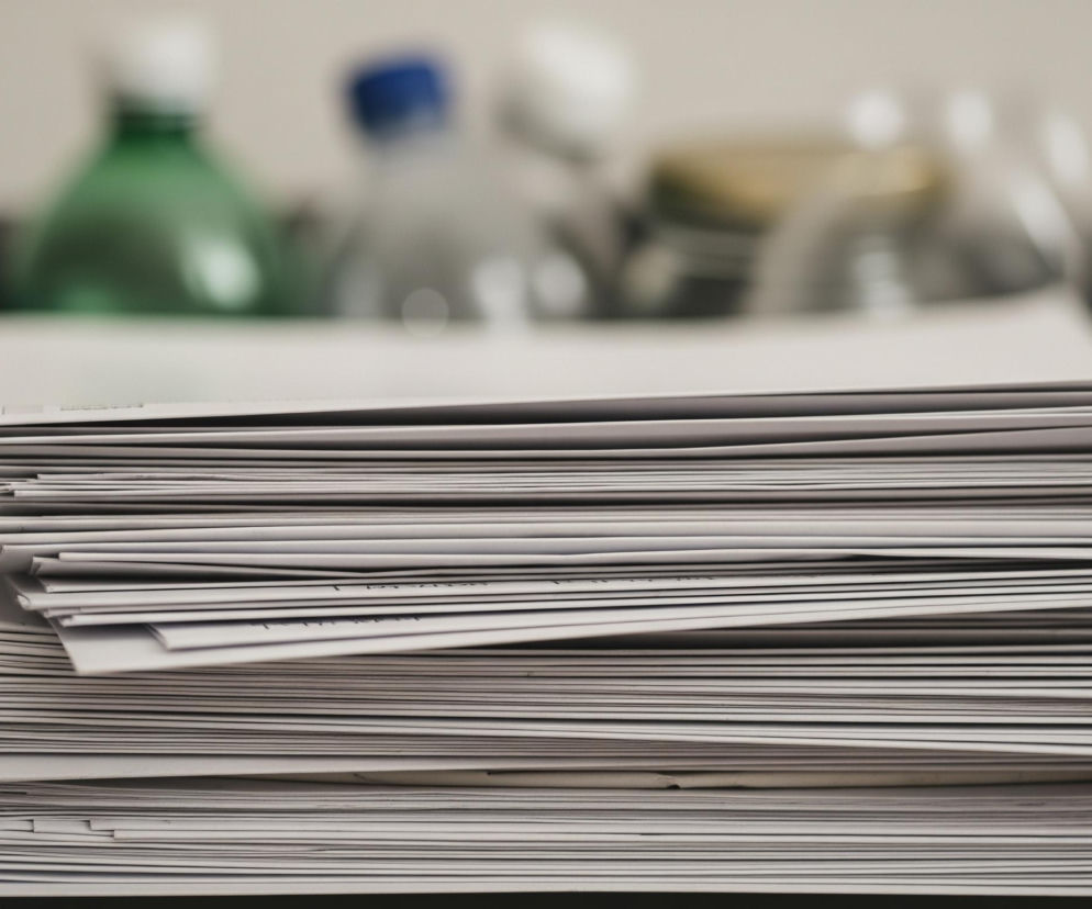 Na pierwszym planie, centralnie, znajduje się duży stos białych kopert lub dokumentów, ułożonych równo i ciasno jeden na drugim. Koperty są w różnych odcieniach bieli i szarości, niektóre z widocznymi zagięciami lub drobnymi uszkodzeniami krawędzi. Stos leży na ciemnoszarym lub czarnym, płaskim przedmiocie, który częściowo wystaje z lewej strony kadru. W tle, rozmyte i nieostre, widać kilka plastikowych butelek – jedną z zieloną nakrętką i zielonkawym odcieniem, drugą z niebieską nakrętką i bezbarwną, oraz dwie inne, również rozmyte, z niewyraźnymi elementami w kolorze żółtym i białym. Tło za butelkami jest jasnoszare i jednolite.