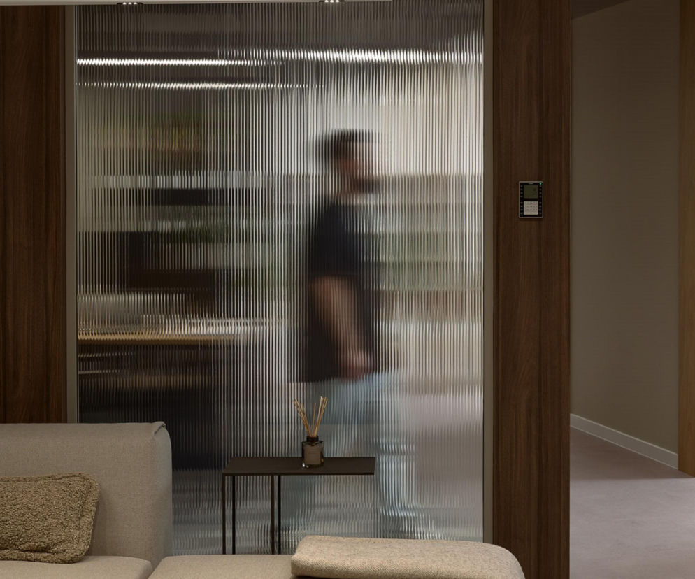 Nowoczesne wnętrze biura CMT Advisory w Poznaniu, z beżową sofą i stolikiem kawowym w centrum uwagi. Widoczny element szklanej ścianki działowej z efektem prążkowania, za którą w tle, delikatnie rozmyty, przesuwa się mężczyzna. Drewniane wykończenia ścian podkreślają elegancję przestrzeni, o której więcej na Architektura Murator Plus.