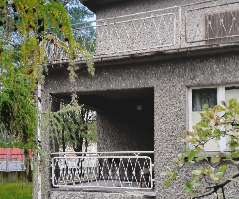 Szary, piętrowy dom w kształcie kostki z lat 70., pokryty tynkiem barankiem, z białymi barierkami balkonowymi i loggią na parterze. Otoczenie zielone z drzewami i niską roślinnością. Dowiedz się więcej o termomodernizacji podobnych budynków na naszym portalu.