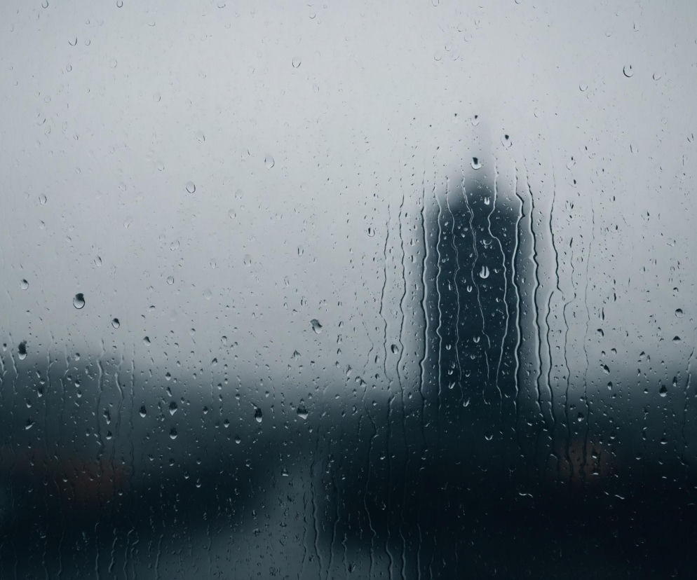 Przednia szyba, pokryta licznymi, różnej wielkości kroplami deszczu, wypełnia cały kadr. Na rozmazanym tle widać ciemnoszarą, smukłą sylwetkę budynku, sprawiającą wrażenie wieżowca, z której po pionowych smugach spływają krople wody. Górna część tła jest jaśniejsza, przyjmując szarobiały odcień, natomiast dolna część jest znacznie ciemniejsza, prawie czarna.