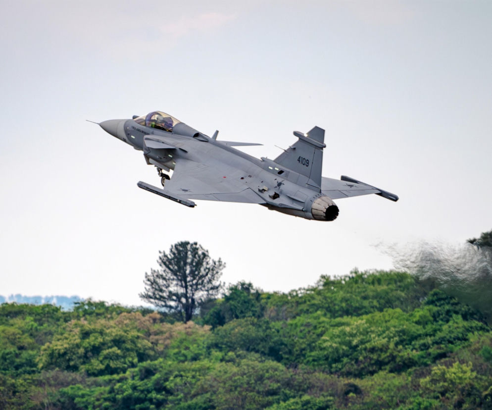 Szary myśliwiec F-39E Gripen podczas startu nad zielonymi drzewami. O sukcesie brazylijskiego przemysłu przeczytasz na SE Portal Obronny.