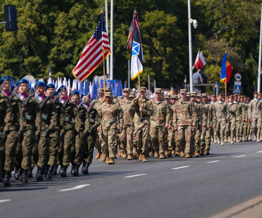 Żołnierze polscy w mundurach galowych z niebieskimi beretami i bronią maszerujący obok żołnierzy amerykańskich w mundurach polowych z flagą USA, co symbolizuje silne poparcie społeczne dla stałej bazy wojsk USA w Polsce, o którym możesz przeczytać na naszym portalu.