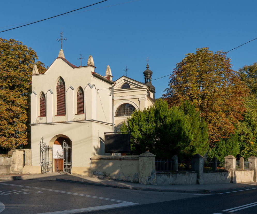 Kościół pw. św. Stanisława Biskupa w Osieku, widoczny od strony ulicy Sandomierskiej, otoczony zielenią i fragmentem muru cmentarnego. Fotografia ukazuje zabytkowy obiekt, który od lat borykał się z problemem uciążliwego ruchu drogowego, o czym przeczytasz na naszym portalu.