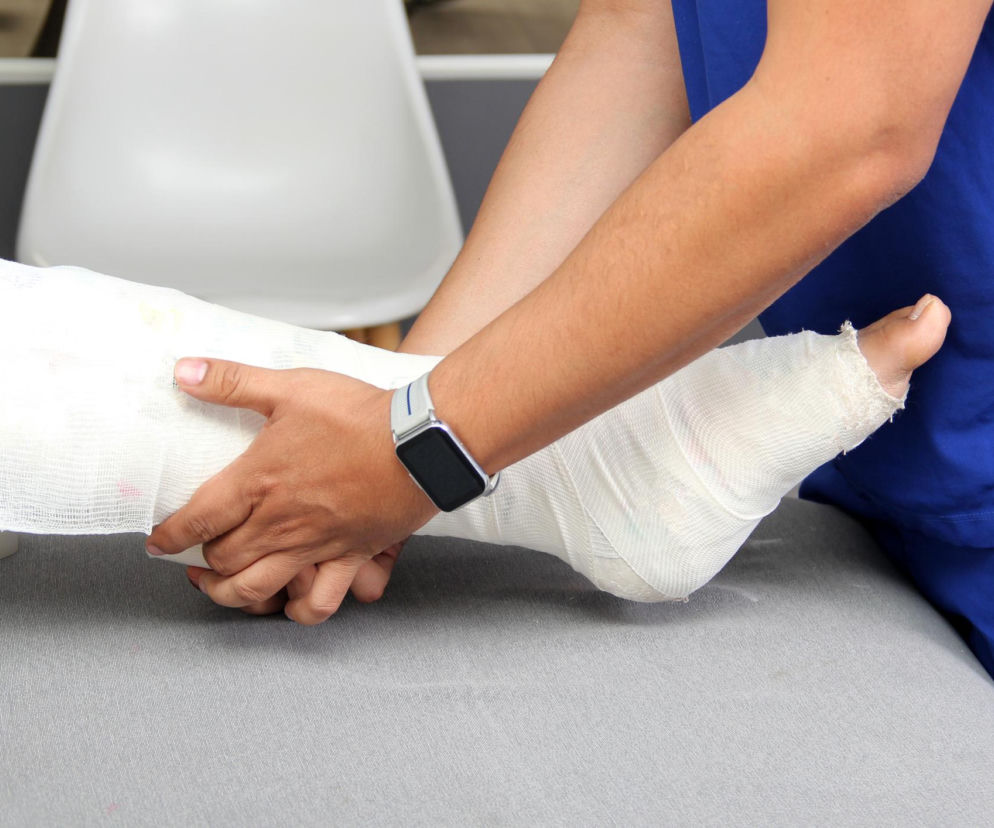 Osoba w niebieskim stroju medycznym delikatnie przytrzymuje zabandażowaną nogę leżącego pacjenta, co wskazuje na pomoc w przypadku urazu. Artykuł Poradnik Zdrowie przestrzega przed nagłym wzrostem liczby złamań i urazów spowodowanych śliskimi chodnikami.