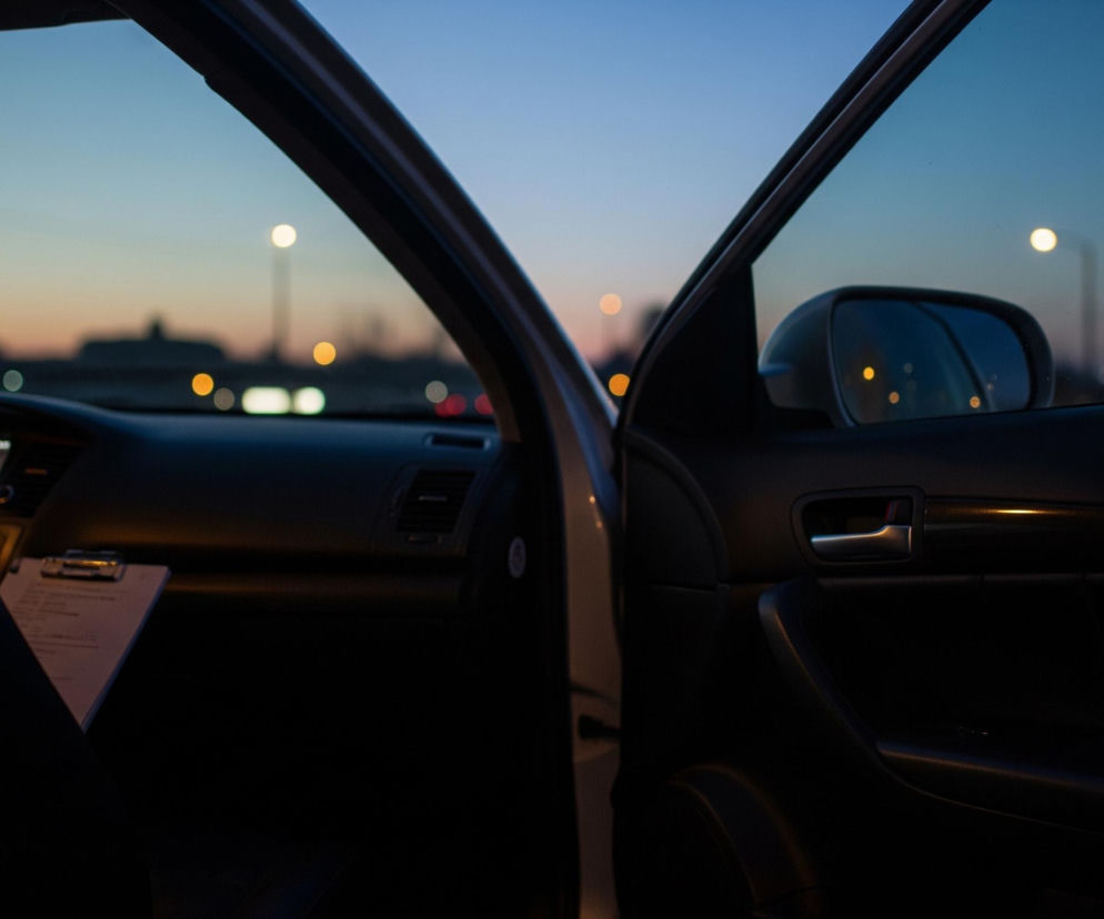 Scena ukazuje wnętrze samochodu, widziane od strony siedzenia pasażera, z otwartymi drzwiami kierowcy i pasażera. Na desce rozdzielczej widoczny jest ekran informacyjny oraz schowek z clipboardem. Na zewnątrz, pod błękitno-pomarańczowym niebem, widać rozmyte światła uliczne i sylwetki budynków, a w lusterku bocznym odbijają się dodatkowe, rozproszone źródła światła. Całość jest skąpana w ciepłym, wieczornym lub porannym świetle.