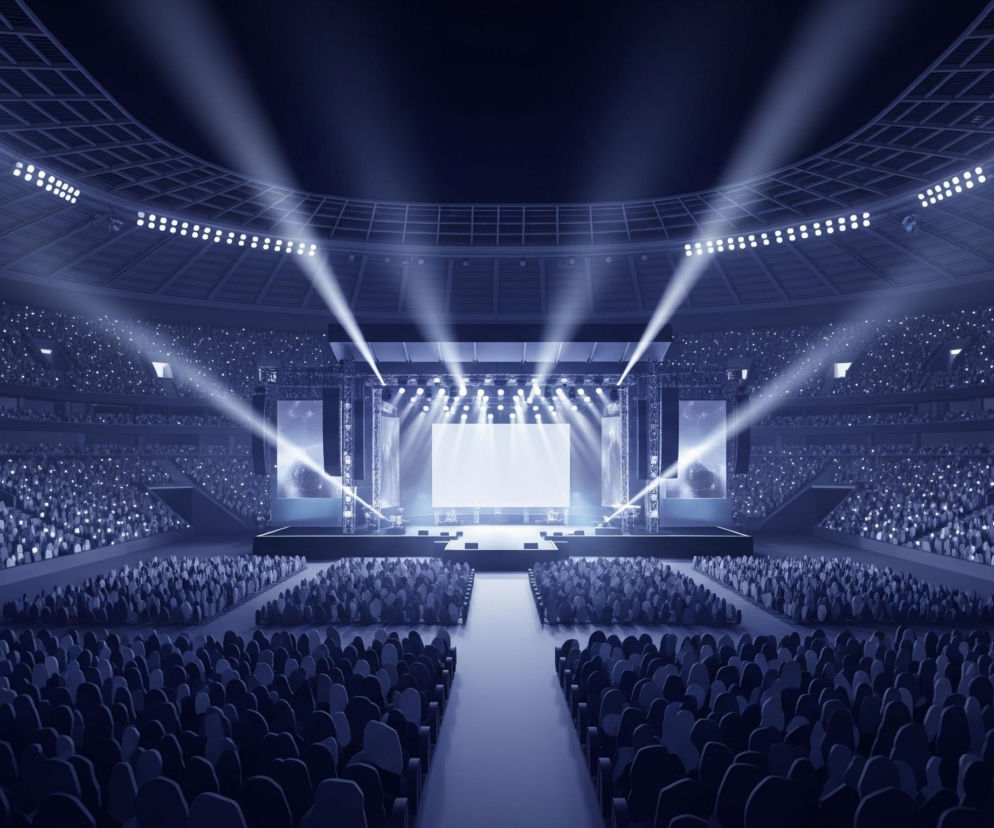 Stadion w ciemnych odcieniach niebieskiego wypełniony jest publicznością z tysiącami podniesionych, świecących telefonów. Scena na pierwszym planie, oświetlona silnymi, białymi snopami światła z reflektorów sufitowych i bocznych, otoczona jest dużymi ekranami wyświetlającymi obrazy. Konstrukcje sceniczne oraz fragmenty dachu stadionu są wyraźnie widoczne w górnej części kadru.