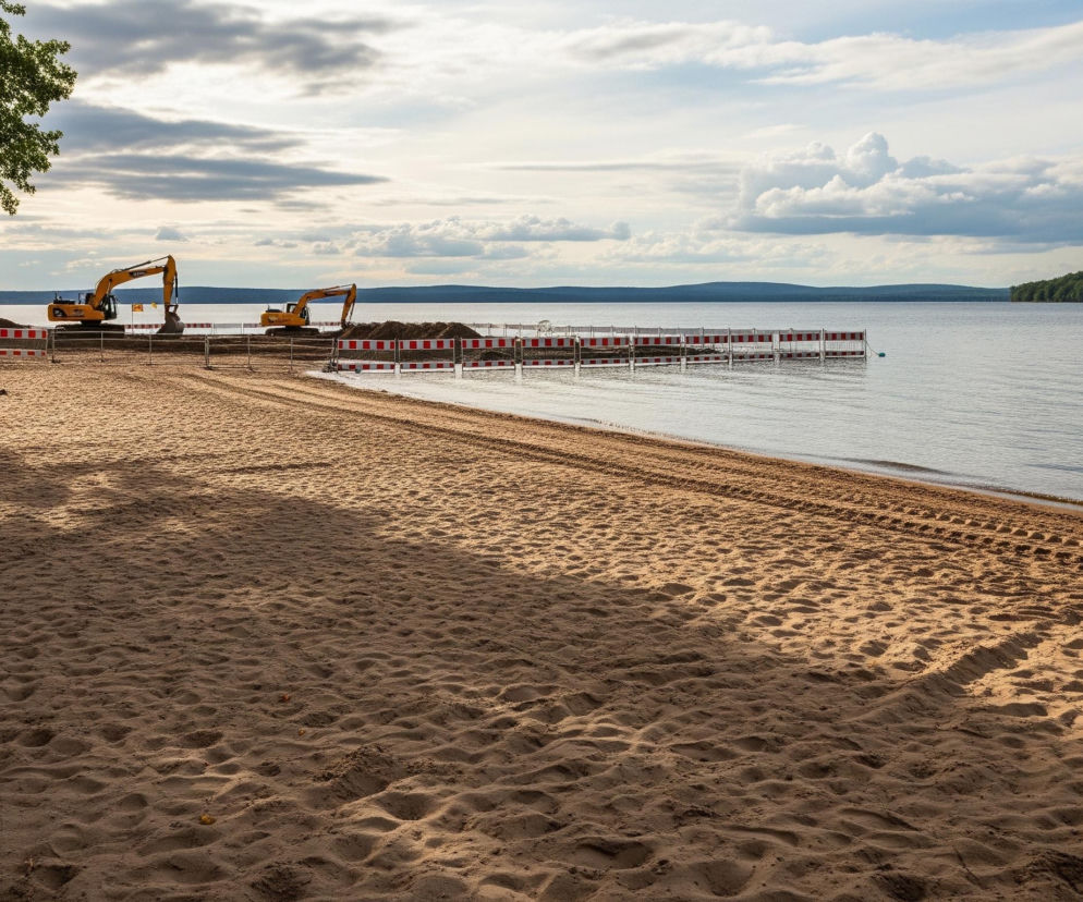 Piaszczysta plaża rozciąga się wzdłuż brzegu jeziora, zajmując większą część pierwszego planu, z ciemnymi cieniami drzew z lewej strony. Na plaży widoczne są ślady opon oraz stosy piasku. Dwa żółte koparki stoją przy wodzie, częściowo ogrodzone biało-czerwonymi barierkami, które biegną wzdłuż brzegu, a następnie wchodzą w głąb jeziora. Po drugiej stronie jeziora, na horyzoncie, rozpościera się ciemnozielony las pod niebem z białymi i szarymi chmurami.