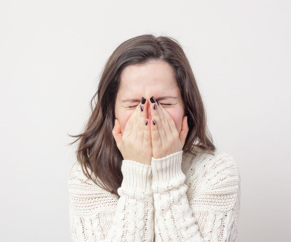 Młoda kobieta z długimi, ciemnymi włosami, w jasnym, dzierganym swetrze, zasłania twarz dłońmi, próbując stłumić kichnięcie, co może prowadzić do poważnych konsekwencji zdrowotnych. Więcej o tym, dlaczego nie wolno wstrzymywać kichania, przeczytasz na Poradnik Zdrowie.