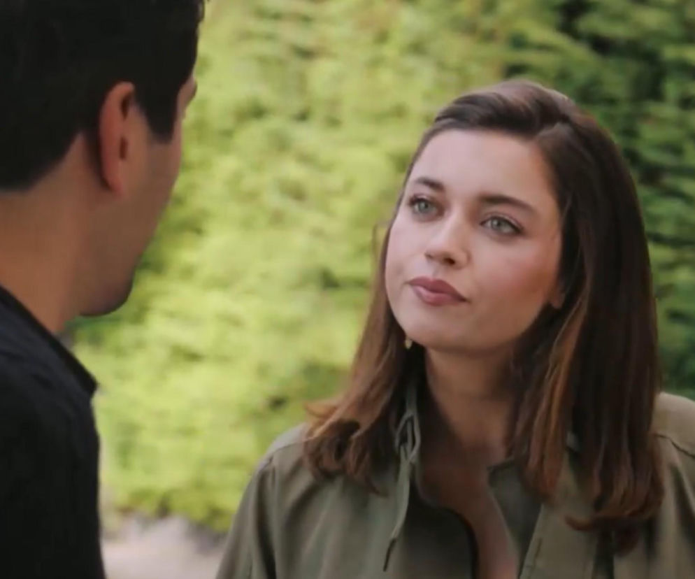 Kobieta z brązowymi włosami i zielonymi oczami, Seyran, patrzy z wyraźnym smutkiem na stojącego tyłem mężczyznę, Ferita, z rozczarowaniem na twarzy, na tle rozmytych zielonych krzewów. Zdjęcie ilustruje trudną konwersację bohaterów serialu Złoty chłopak, o której więcej przeczytasz na Super Seriale.