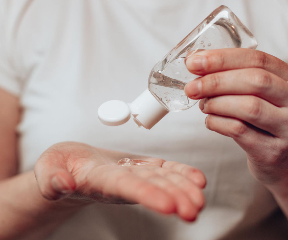 Dłoń wylewająca żel antybakteryjny z małej przezroczystej butelki z białą zakrętką. Kropla bezbarwnego żelu spływa na otwartą dłoń. To nawiązanie do artykułu na Poradnik Zdrowie o tym, czy żel antybakteryjny może szkodzić skórze.