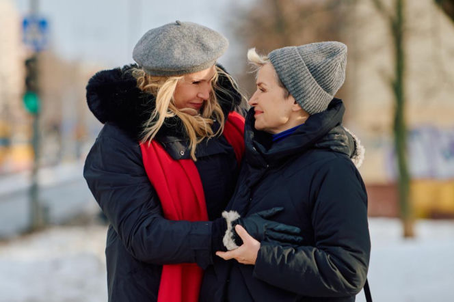 Dwie kobiety w zimowych ubraniach, szarych czapkach i ciemnych kurtkach, delikatnie się obejmują, patrząc sobie w oczy na tle zaśnieżonego parku. Scena sugeruje wsparcie w trudnych chwilach, co doskonale pasuje do dramatycznych wydarzeń z serialu Na Wspólnej, o których można przeczytać na Super Seriale.