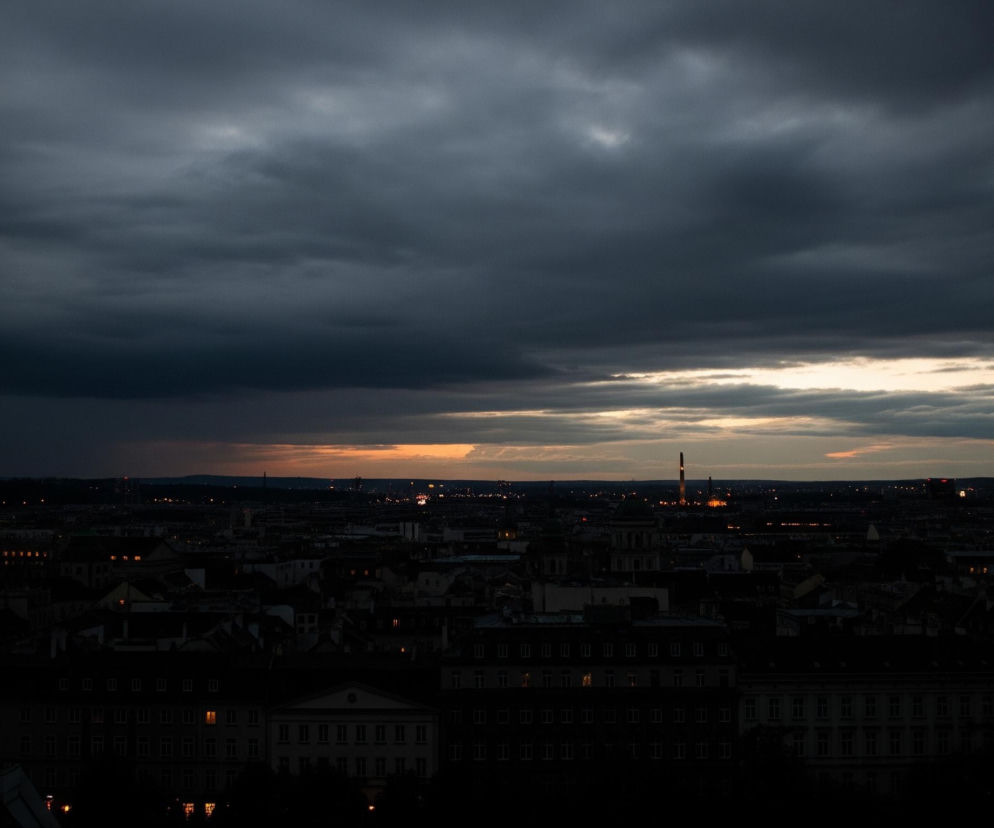 Ciemne, burzowe chmury dominują niebo, zajmując większą część górnej połowy kadru, z nielicznymi jasnymi smugami pomarańczowego i jasnobeżowego światła widocznymi nisko nad horyzontem. Poniżej rozciąga się miejski krajobraz ukazany w nocnym świetle, z rzędami budynków i kilkoma świecącymi oknami, tworzącymi jasne, żółte punkty. Horyzont jest usiany licznymi, drobnymi światłami, które zlewają się w linie, a w oddali widać wysoką, ciemną strukturę przypominającą komin, oświetloną u szczytu.