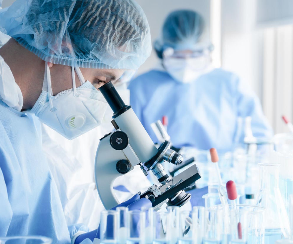 Naukowiec w kombinezonie ochronnym i masce FFP3, pochylony nad mikroskopem w laboratorium, z rzędami probówek i kolb z niebieską cieczą. W tle rozmyta postać drugiego naukowca. O nowej szczepionce na hantawirusa przeczytasz na Poradniku Zdrowie.