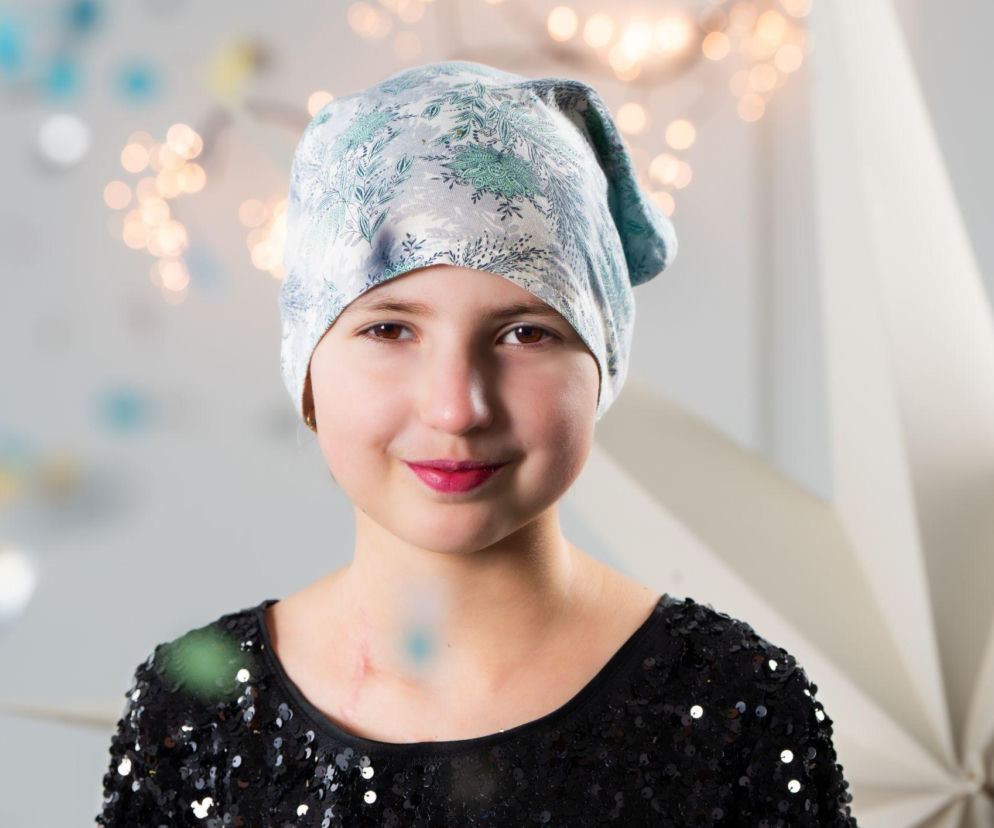Dziesięcioletnia Emilka Wójcik uśmiecha się do obiektywu, nosząc wzorzystą chustę na głowie i błyszczącą czarną sukienkę. Za nią widoczne są rozmyte światła i dekoracja w kształcie gwiazdy, co nadaje zdjęciu radosny i świąteczny nastrój, symbolizujący nadzieję. Na portalu Poradnik Zdrowie można przeczytać o jej akcji Postanawiam Pomagać.