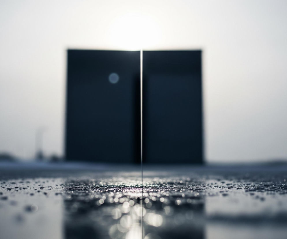 Na obrazie dominuje ciemna, minimalistyczna kompozycja. Na pierwszym planie widoczna jest powierzchnia pokryta licznymi, błyszczącymi kroplami wody, z efektem bokeh, co sugeruje odbicie światła. W tle, centralnie umiejscowiony jest prostokątny, ciemny obiekt podzielony pionową, jasną linią światła, tworząc wrażenie otwartych drzwi lub szczeliny. Całość utrzymana jest w chłodnych odcieniach szarości, bieli i granatu.