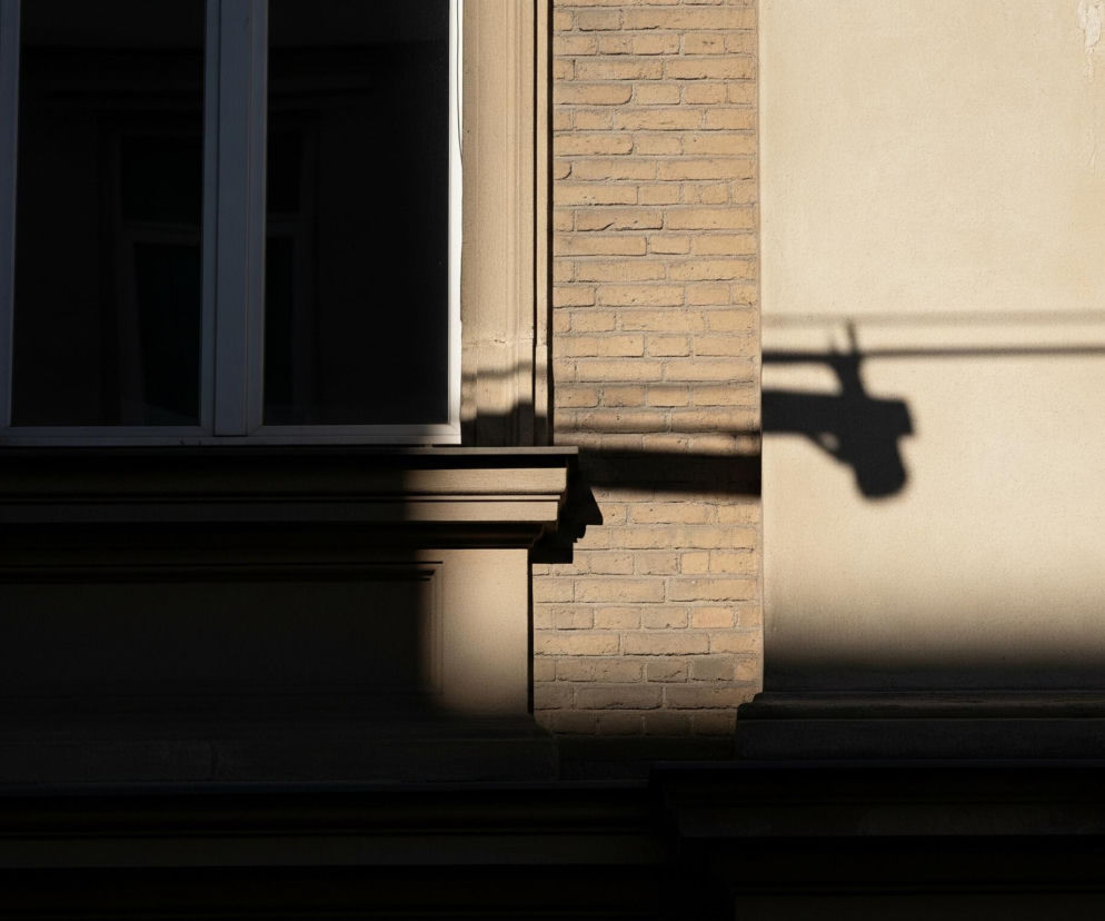 Fragment fasady budynku z dominującym podziałem na światło i cień. Po lewej stronie znajduje się pionowe okno z białą ramą, którego szklana powierzchnia jest ciemna i odbija niewiele światła. Poniżej okna widoczny jest ciemny, ozdobny gzyms, również w cieniu. Bezpośrednio na prawo od okna rozciąga się mur z żółtej cegły, częściowo oświetlony ukośnym światłem, rzucającym długie, cienkie cienie. Prawa część obrazu to jasnobeżowa ściana pokryta tynkiem, silnie oświetlona słońcem, na której widoczne są czarne, rozmyte cienie, przypominające sylwetki przewodów lub gałęzi.