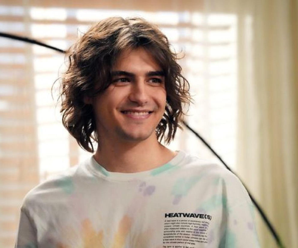 Uśmiechnięty Jacek Tyszkiewicz jako Romeo w serialu Barwy szczęścia, ubrany w białą koszulkę w barwne plamy, patrzący w prawo. Obok fragment rośliny i jasne tło. Więcej o losach Romea przeczytasz na portalu Super Seriale.