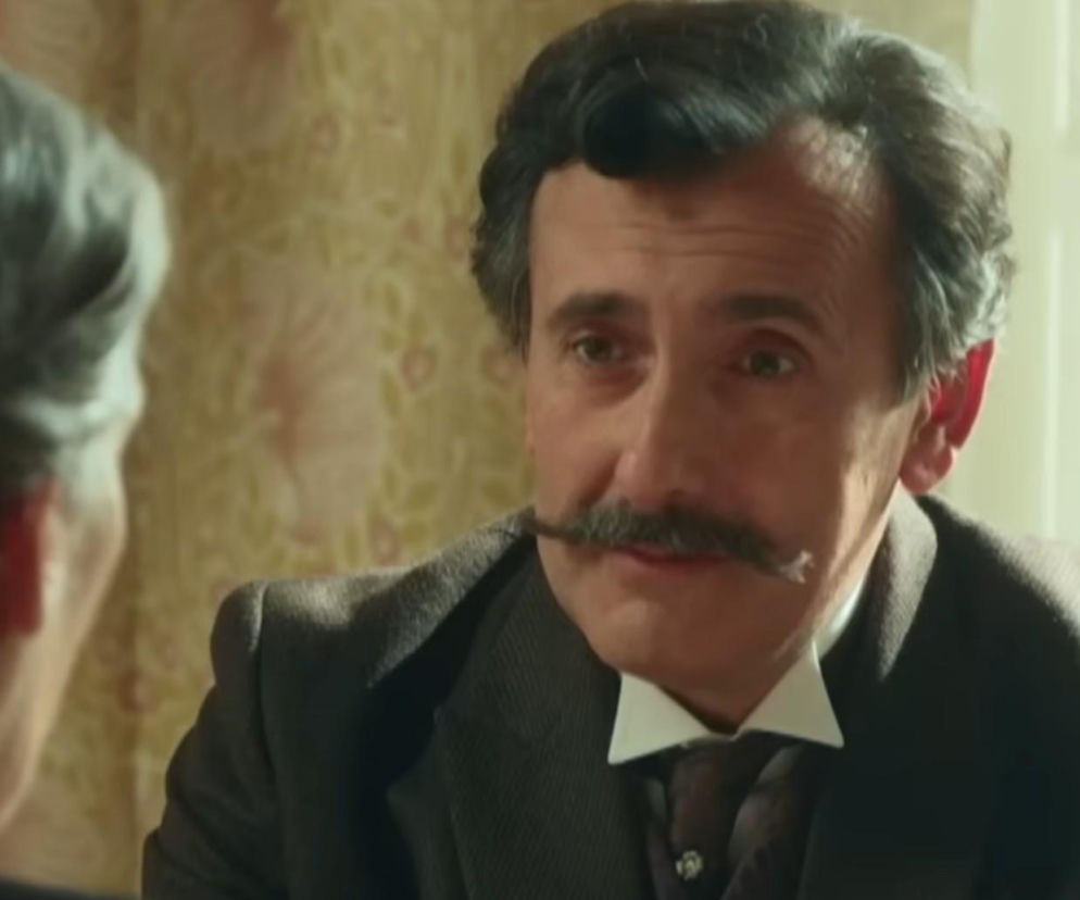 Mężczyzna o siwych włosach i bujnych wąsach w ciemnym garniturze i białej koszuli z krawatem, patrzący z powagą na niewidoczną rozmówczynię. To scena z serialu La Promesa, gdzie Ayala zaskakuje Petrę wyznaniem. Więcej o ich relacji i intrygach przeczytasz na Super Seriale.