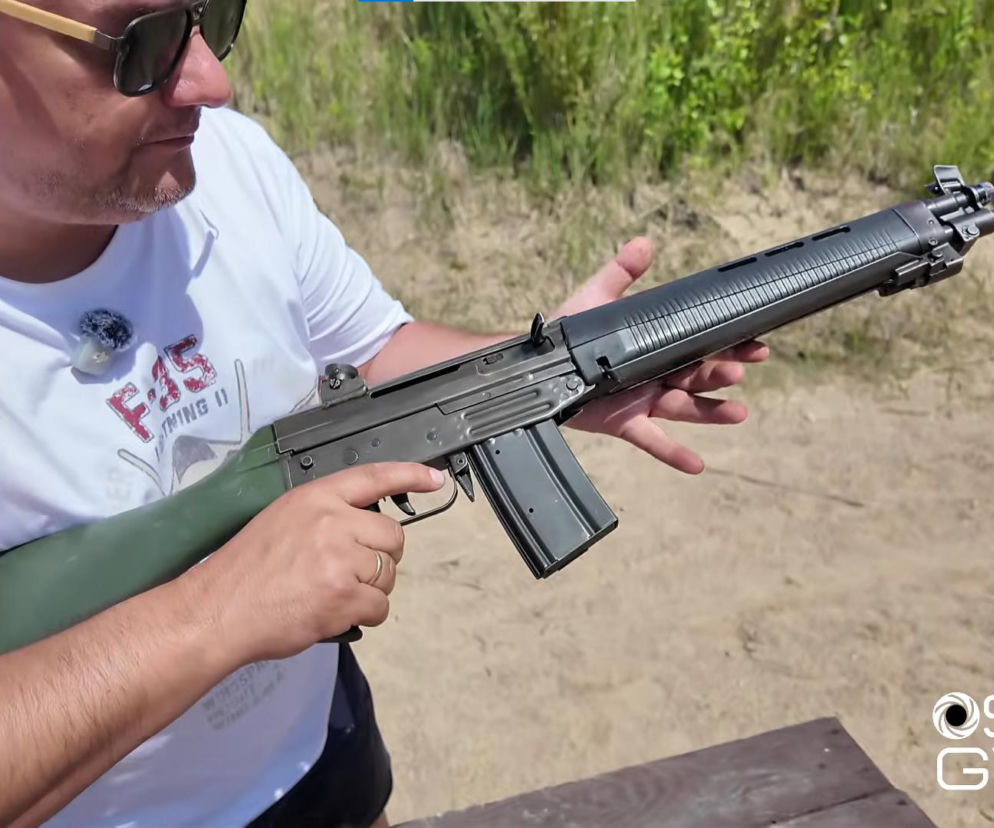 Mężczyzna w okularach i białej koszulce trzyma karabinek SIG SG 540 z zieloną kolbą, prezentując jego budowę. Broń, ważny element ewolucji szwajcarskiej myśli technicznej, to zapomniany ojciec legendy SG 550, o czym przeczytasz więcej na naszym portalu.