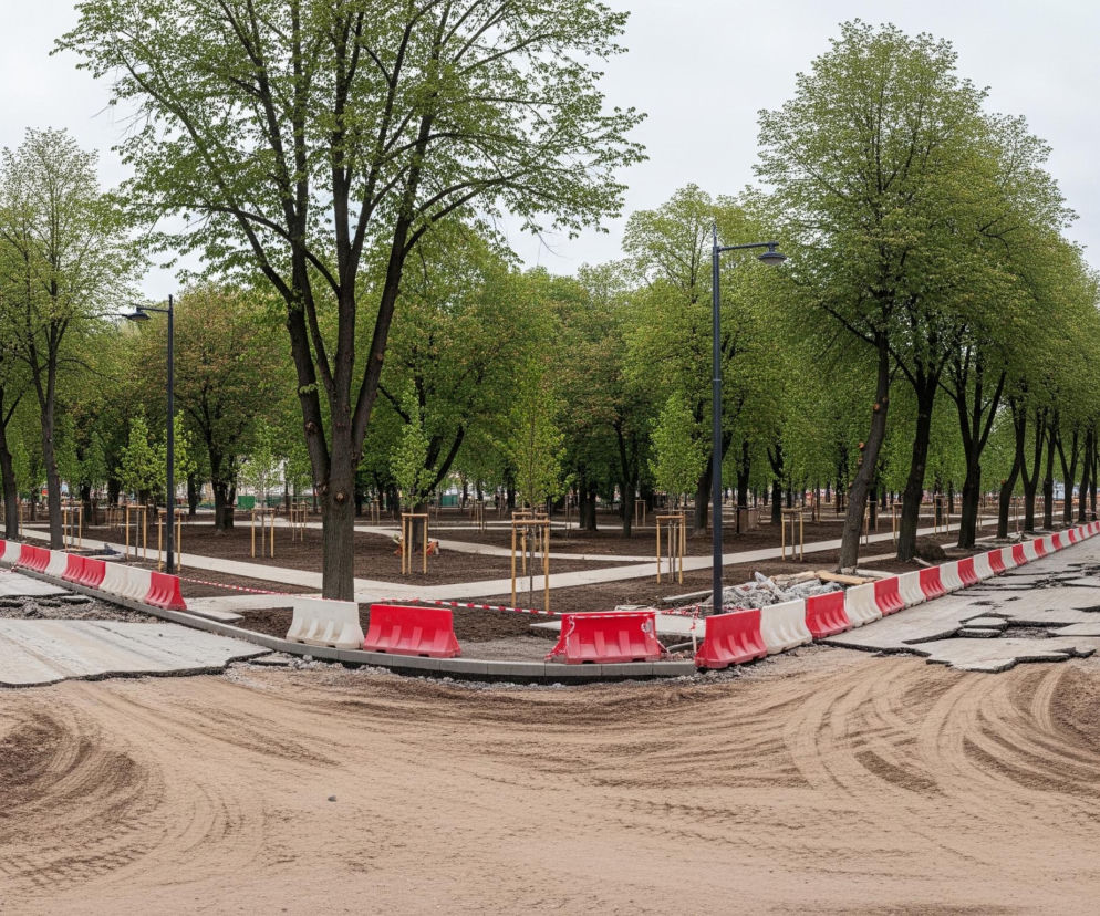 Przestrzeń miejska w trakcie remontu, z widokiem na skrzyżowanie rozkopanych ulic, które rozdzielają się w lewo i prawo, otoczone alejami drzew o jasnozielonych liściach. Nawierzchnia dróg jest rozebrana, odsłaniając warstwy gruzu i piachu, a pośrodku znajduje się piaszczysty obszar z widocznymi śladami opon. Obie strony rozkopanych ulic są zabezpieczone biało-czerwonymi barierami budowlanymi, a za nimi rozciąga się park z nowo posadzonymi drzewkami i ścieżkami, z szarym, zachmurzonym niebem w tle.
