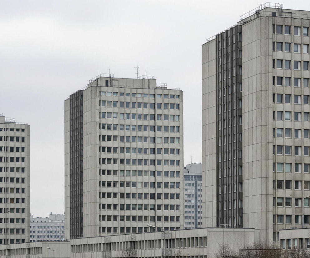 Trzy wysokie, prostopadłościenne bloki z szarego betonu dominują obraz, każdy z nich posiada liczne, identycznie ułożone okna w rzędach i kolumnach. Na dachach budynków widoczne są anteny i metalowe barierki, a na bocznych elewacjach niektórych z nich ciemniejsze, pionowe pasy z mniejszymi oknami. Na pierwszym planie, u podstawy budynków, rozciąga się niższy, podłużny budynek również z rzędami okien, przed nim rosną bezlistne drzewa, a w tle widać inne, mniej wyraźne budynki. Całość otacza jasne, zachmurzone niebo.