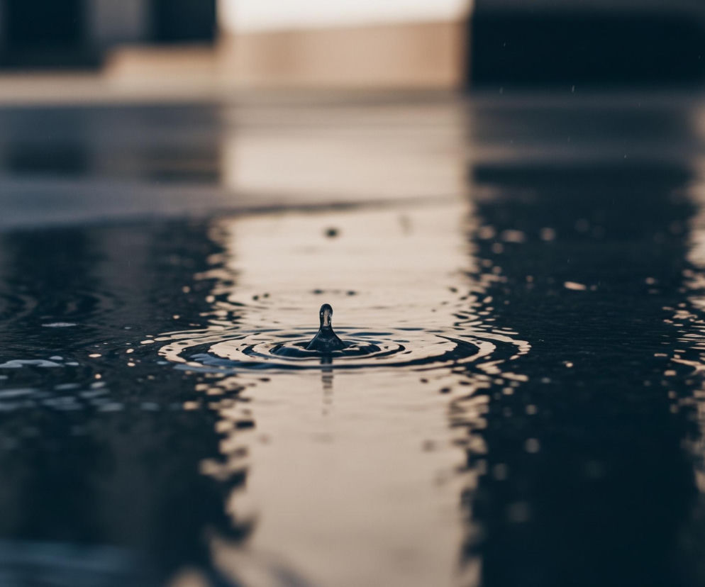 Kropla wody wznosi się nad ciemną powierzchnią wody, tworząc koncentryczne kręgi fal rozchodzące się na boki. Woda jest ciemnoniebieska, odbijając światło w tonacji beżowo-brązowej, zwłaszcza w tle, gdzie zamazane kontury sugerują odległe obiekty. Całość sprawia wrażenie spokojnej, lecz dynamicznej sceny, z widocznymi drobinkami wody w powietrzu.