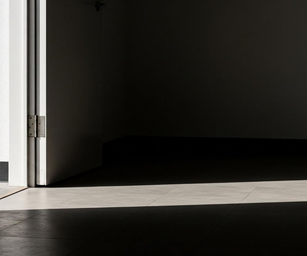 Wnętrze pomieszczenia z uchylonymi drzwiami, tworzące silny kontrast między jasnymi a ciemnymi obszarami. Po lewej stronie dominuje ciemna, częściowo widoczna powierzchnia, prawdopodobnie wewnętrzna strona drzwi, a obok niej jaśniejsza, biała futryna z widocznym, srebrnym zawiasem. Dalej rozpościera się ciemne pomieszczenie, z fragmentem podłogi wyłożonej jasnoszarymi płytkami, na którą pada pozioma smuga ostrego światła, rozciągająca się od futryny w głąb kadru.