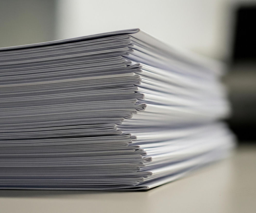 Duży stos białych kartek papieru, ciasno ułożonych jedna na drugiej, dominuje środek obrazu, zajmując prawie całą lewą połowę kadru. Brzegi kartek, widoczne z boku, tworzą regularne, poziome linie w odcieniach szarości i bieli. Głębia ostrości skupia się na pierwszym planie, podczas gdy tło jest rozmyte, z jasnoszarym odcieniem biurka w dolnej części i ciemniejszymi, niewyraźnymi elementami w górnym prawym rogu.
