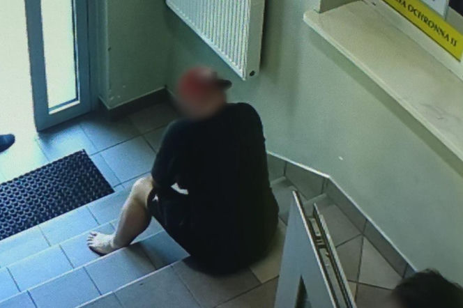 Mężczyzna z zakrytą twarzą i w czapce siedzi boso na schodach w budynku komendy policji w Mielcu, co odnosi się do kuriozalnej kradzieży łańcuszka i ucieczki złodzieja. O sprawie przeczytasz więcej na naszym portalu.